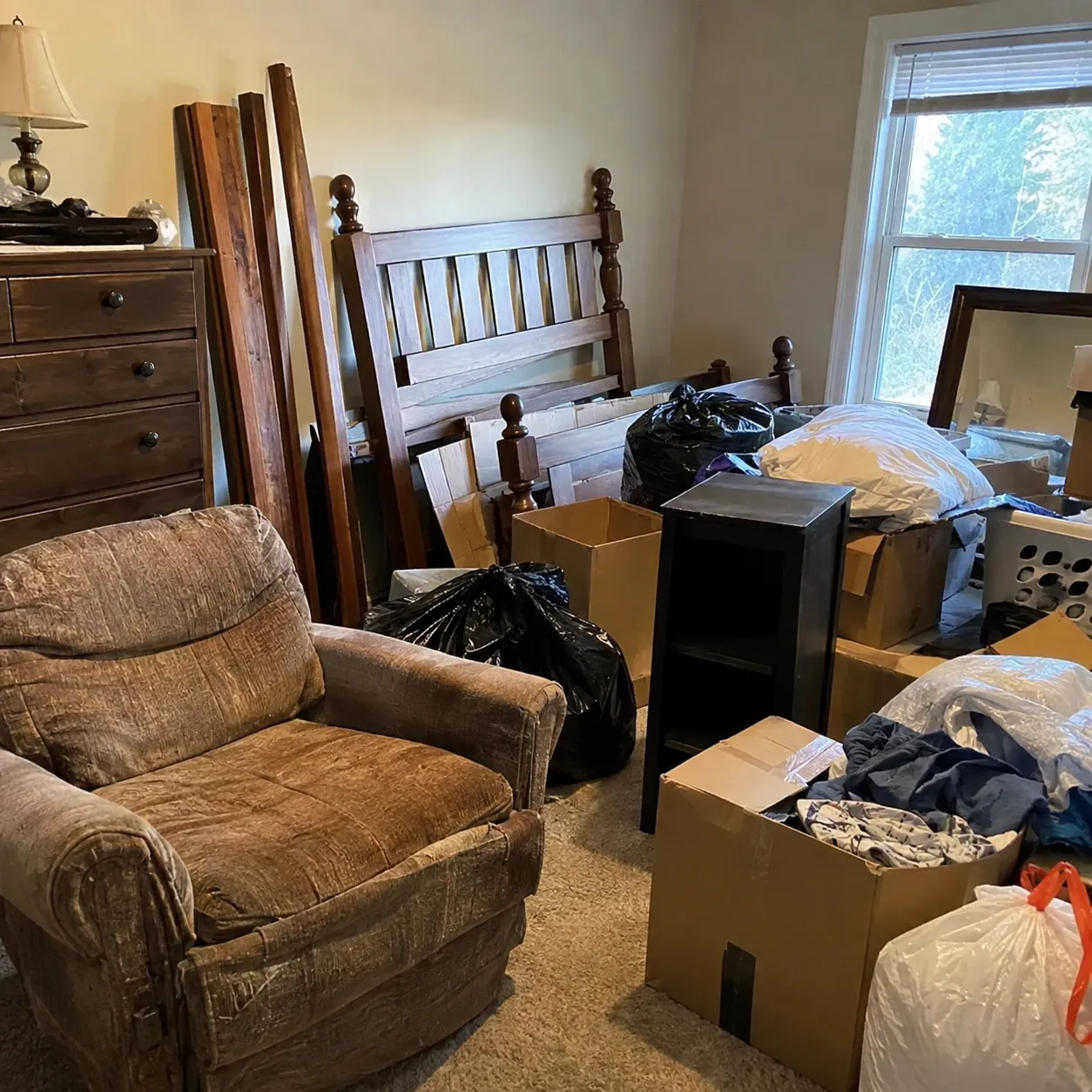 A bedroom filled with a bed frame, armchair, boxes and bagged belongings awaiting removal